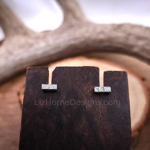 May include: A pair of silver stud earrings with a textured rectangular design. The earrings are displayed on a wooden stand.