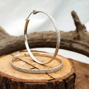 May include: A pair of silver hoop earrings featuring a textured, intricate floral pattern on the outer surface. The earrings are circular with a thin profile and a hinged closure mechanism, showcasing a metallic finish with detailed engravings.