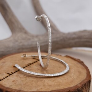 May include: A pair of silver hoop earrings with a textured finish. The earrings are displayed on a wooden surface.
