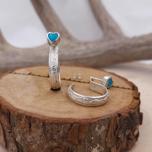 May include: A pair of silver hoop earrings with turquoise heart accents. The earrings are on a wooden surface.