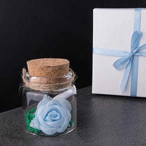 Puede incluir: Un pequeño tarro de cristal con tapón de corcho y detalles de cuerda contiene una rosa de tela azul claro. Una caja de regalo blanca atada con una cinta azul claro está al lado del tarro. El fondo es negro y gris.