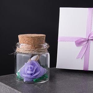 Puede incluir: Una pequeña botella de vidrio con tapón de corcho, hilo y una rosa morada en su interior. Una caja de regalo blanca con una cinta y lazo lila está junto a la botella. El fondo es negro y los objetos están sobre una superficie gris.