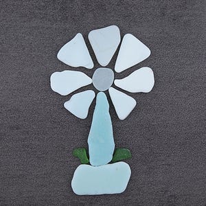 Puede incluir: Un diseño floral hecho con piezas de vidrio de mar en tonos blanco, azul claro y verde. La flor tiene un centro gris y está dispuesta sobre un fondo gris oscuro. TheSeaDreamers.etsy.com está impreso en la parte inferior derecha.