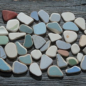 Puede incluir: Una colección de azulejos de cerámica lisos, coloridos y de formas únicas en tonos de azul, verde, blanco y marrón. Los azulejos están dispuestos en un patrón circular sobre una superficie de madera desgastada.