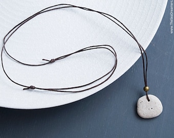 May include: A handmade necklace featuring a small, light gray stone pendant. The stone is attached to a dark brown cord with adjustable knots. The necklace is displayed on a white surface with a dark blue background. The website address is visible in the top right corner.