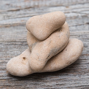 Può includere: Una pila di ciottoli di fiume lisci e color beige chiaro, disposti in una formazione equilibrata. Le pietre hanno una forma arrotondata e una consistenza naturale e usurata. Lo sfondo è una superficie di legno invecchiato.