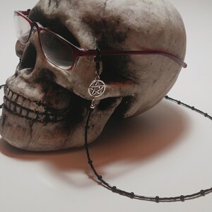 May include: A skull wearing burgundy-framed glasses with a black beaded eyeglass chain. A silver pentagram charm hangs from the glasses. The skull is off-white and sits against a white background.