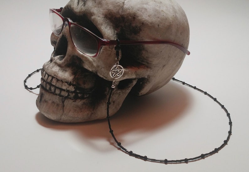 May include: A skull wearing burgundy-framed glasses with a black beaded eyeglass chain. A silver pentagram charm hangs from the glasses. The skull is off-white and sits against a white background.