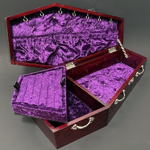 May include: A dark wood coffin-shaped jewelry box with a hinged lid, lined in deep purple velvet. The interior features silver-toned metal hooks along the top edge, a removable ring tray insert, and silver-toned metal handles on the exterior sides.