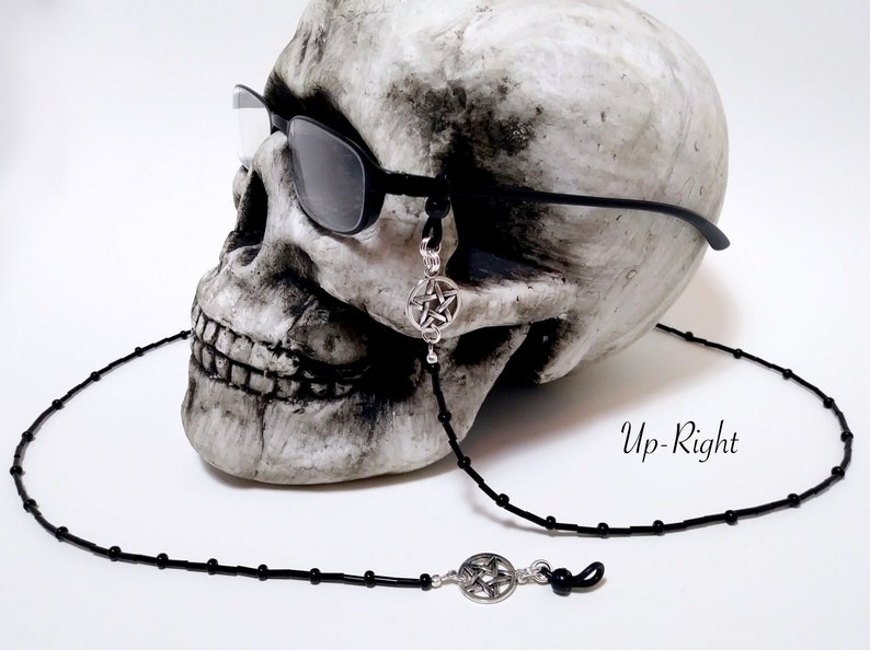May include: Black eyeglasses with a black beaded chain and a silver pentagram charm. The glasses are resting on a skull. The text "Up-Right" is in the lower right corner.