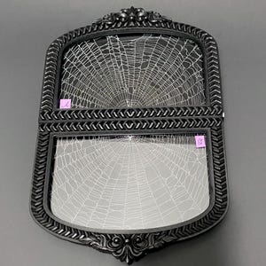 May include: A black double-frame with ornate carved edges and a chevron-patterned center divider. Each of the two glass sections features a white spiderweb design. Small pink labels marked with the letters A and B are attached to the glass.