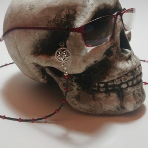 May include: A skull wearing red-framed sunglasses with a black beaded eyeglass chain. A silver pentagram charm hangs from the glasses. The chain has red beads spaced along its length.