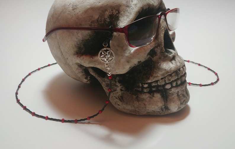 May include: A skull wearing red-framed sunglasses with a black beaded eyeglass chain. A silver pentagram charm hangs from the glasses. The chain has red beads spaced along its length.