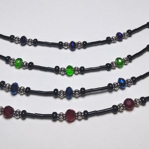 May include: Four beaded necklaces with black cords and silver-colored accents. Each necklace features a central faceted bead in different colors: purple, green, blue, and red. The necklaces are designed for the neck.