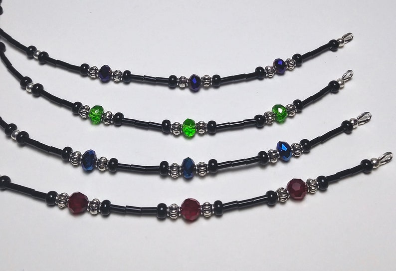 May include: Four beaded necklaces with black cords and silver-colored accents. Each necklace features a central faceted bead in different colors: purple, green, blue, and red. The necklaces are designed for the neck.