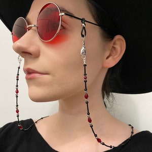 May include: A black beaded glasses chain with red gemstone accents. The chain has a silver clasp and is worn by a person with a black hat and red sunglasses.