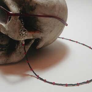 May include: A black beaded eyeglass chain with red accents and a silver pentagram charm. The chain is attached to red-framed sunglasses resting on a skull. The chain is designed to hold glasses.