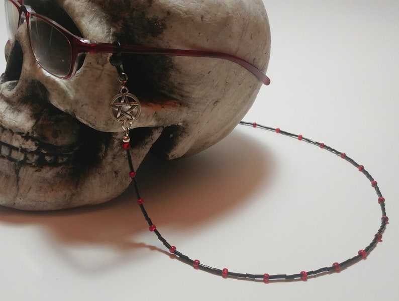 May include: A black beaded eyeglass chain with red accents and a silver pentagram charm. The chain is attached to red-framed sunglasses resting on a skull. The chain is designed to hold glasses.