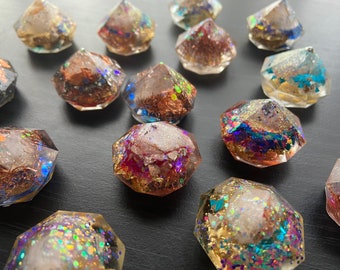 May include: A collection of faceted resin objects with embedded metallic flakes and iridescent glitter. The objects are various colors, including gold, copper, and blue, and are arranged on a dark surface. The overall effect is sparkly and decorative.