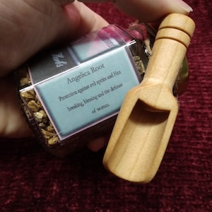 May include: A small glass jar filled with dried herbs and a wooden scoop. The jar has a label that reads "Angelica Root Protection against evil spirits and Hex breaking, blessing and the defense of women."