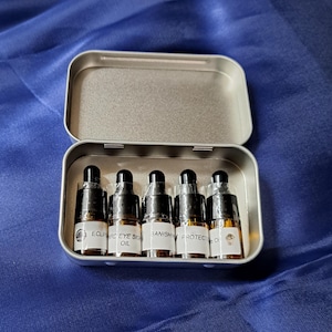 May include: A silver tin box with six small glass bottles containing essential oils. The bottles are labeled with the words "Eclipse", "Eye Serum", "Banish", "Protect", and "Oil".