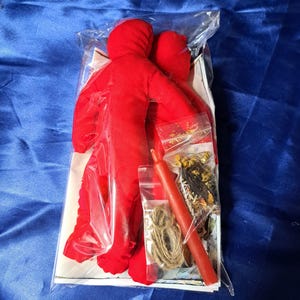 May include: A red doll with a hood, packaged with a red candle, twine, and small bags of herbs and spices. The items are in a clear plastic bag, set against a blue background.
