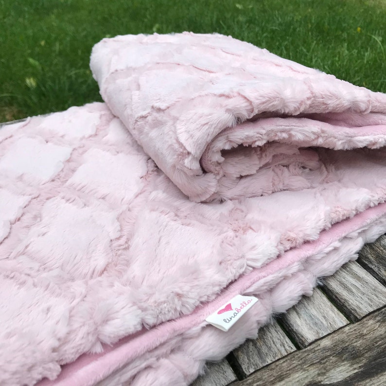 May include: A soft pink plush blanket with a heart-shaped pattern. The blanket is folded in half and has a small tag that says "lina bella".