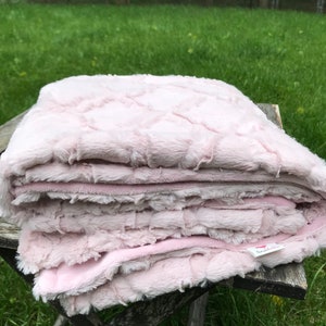 May include: A soft pink faux fur blanket folded on a wooden table. The blanket has a textured pattern and a small white tag with the word "lunaella" on it.