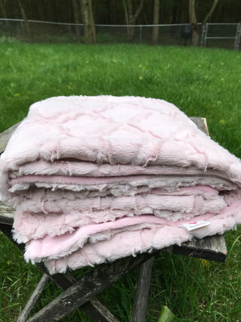 May include: A soft pink faux fur blanket folded on a wooden table. The blanket has a textured pattern and a small white tag with the word "lunaella" on it.