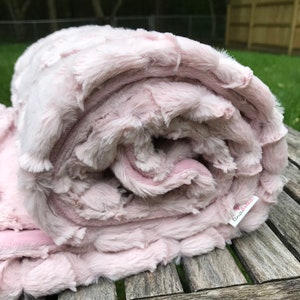May include: A soft, pink, plush blanket rolled up on a wooden surface. The blanket has a textured, fluffy surface and a label that says "linabell".