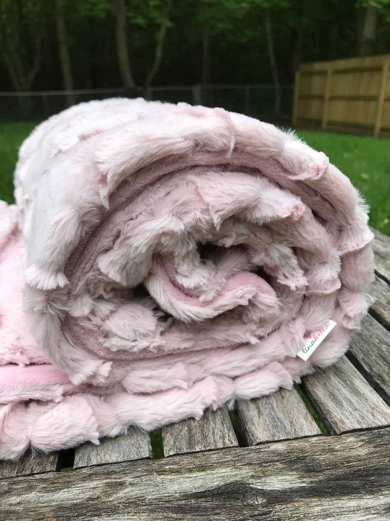 May include: A soft, pink, plush blanket rolled up on a wooden surface. The blanket has a textured, fluffy surface and a label that says "linabell".