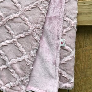 May include: A soft, pink, plush blanket with a geometric pattern on one side and a solid color on the other. The blanket has a small tag with the text "Love Bella's" on it.