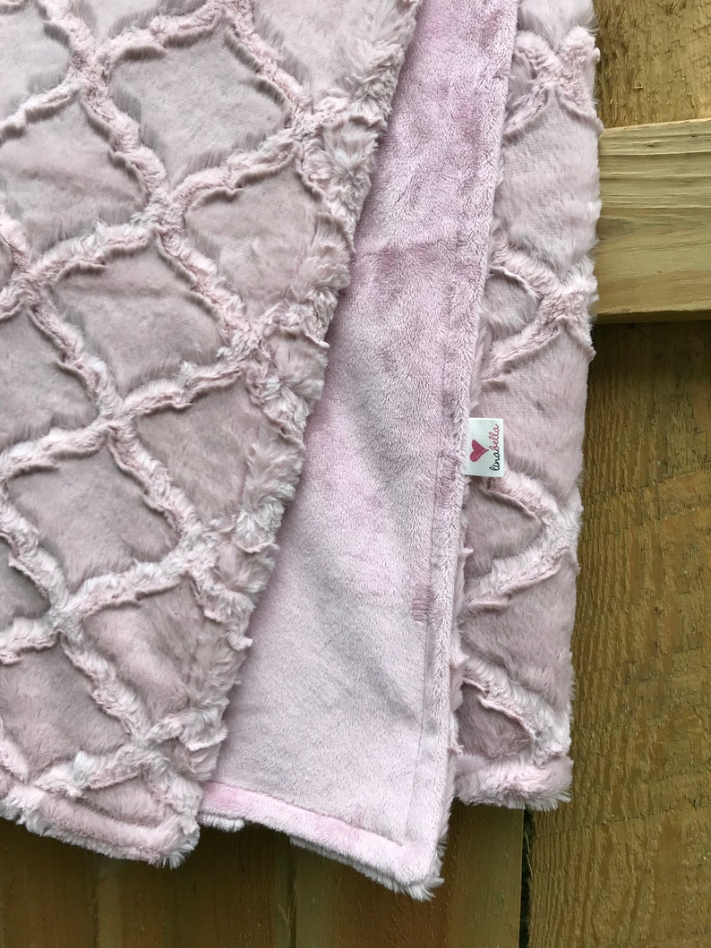 May include: A soft, pink, plush blanket with a geometric pattern on one side and a solid color on the other. The blanket has a small tag with the text "Love Bella's" on it.