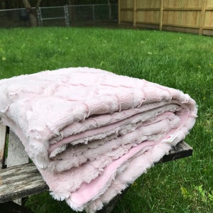 May include: A soft, pink, faux fur blanket folded on a wooden table. The blanket has a textured, quilted pattern.