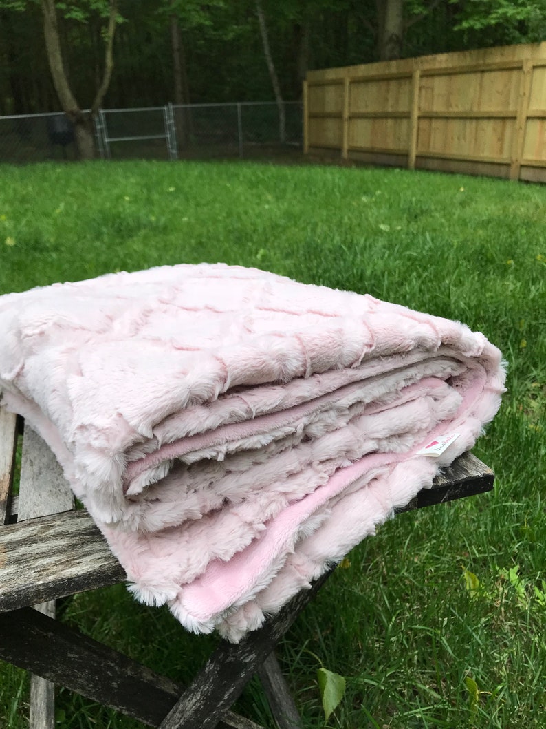 May include: A soft, pink, faux fur blanket folded on a wooden table. The blanket has a textured, quilted pattern.