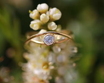 May include: A delicate gold ring with a single round diamond set in a bezel setting. The ring is worn on a finger and is surrounded by white flowers.