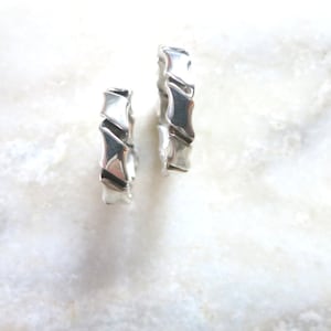 Op de afbeelding: Twee zilveren ringen met een uniek, geometrisch ontwerp. De ringen zijn gemaakt van glad, gepolijst metaal en hebben een licht getextureerd oppervlak.