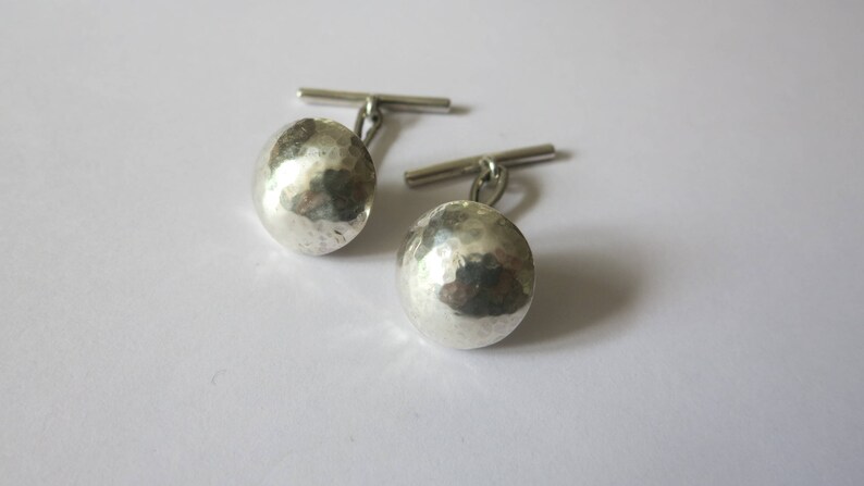 May include: A pair of silver cufflinks with a hammered, textured finish. The cufflinks have a simple, classic design.