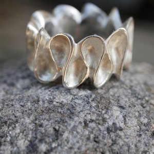 K&ouml;nnte beinhalten: Ein silberner Ring mit einem einzigartigen Design, das aus mehreren abgerundeten, tropfenf&ouml;rmigen Elementen besteht. Der Ring liegt auf einem strukturierten grauen Stein.