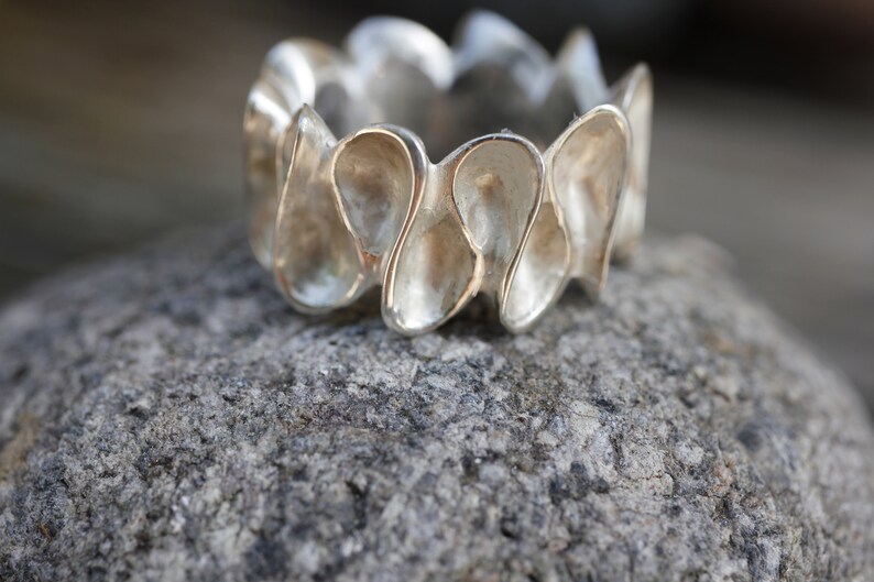 K&ouml;nnte beinhalten: Ein silberner Ring mit einem einzigartigen Design, das aus mehreren abgerundeten, tropfenf&ouml;rmigen Elementen besteht. Der Ring liegt auf einem strukturierten grauen Stein.