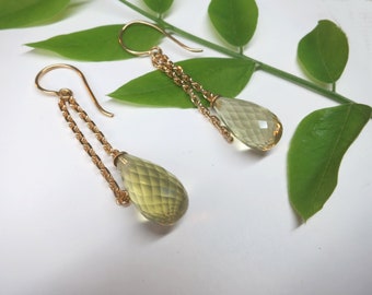 May include: A pair of gold earrings with a teardrop-shaped light green gemstone dangling from each earring. The earrings are on a white background with green leaves.
