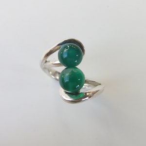 Pode incluir: Um anel de prata com duas contas de pedras preciosas verdes. O anel tem um design único e retorcido.