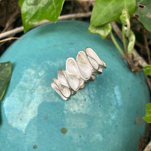 K&ouml;nnte beinhalten: Ein silberner Ring mit einem einzigartigen Design aus mehreren &uuml;berlappenden, gekr&uuml;mmten und strukturierten Teilen. Der Ring wird auf einer t&uuml;rkisfarbenen Oberfl&auml;che gezeigt.