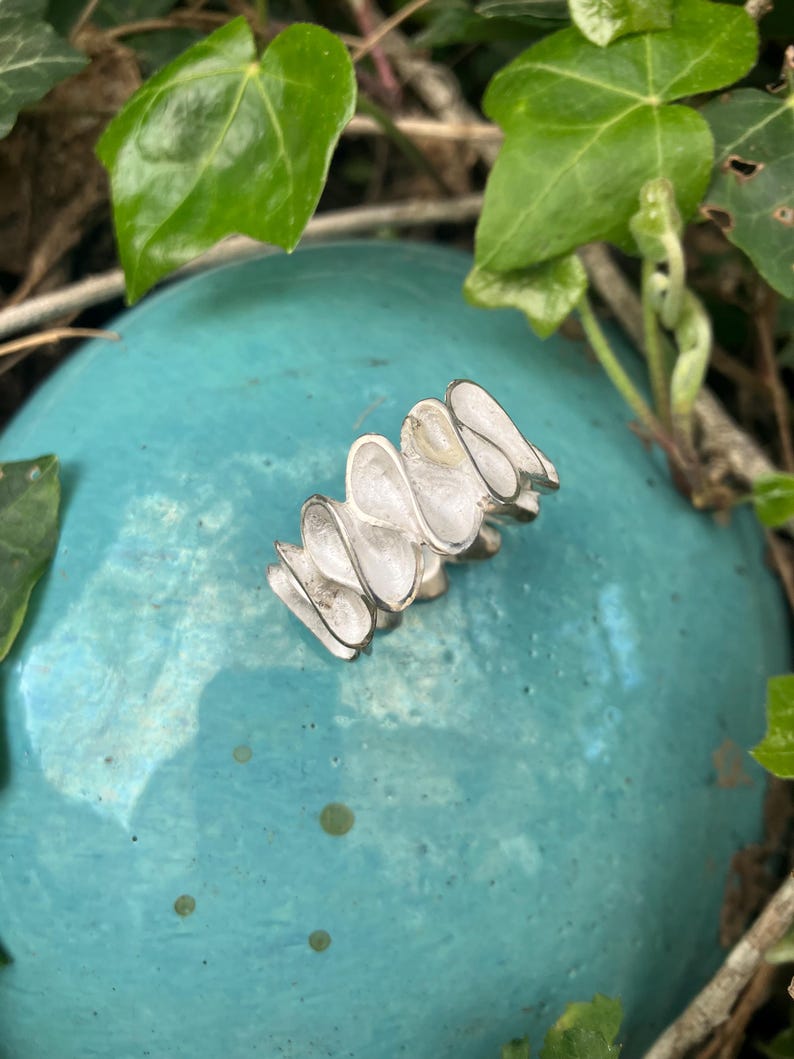 K&ouml;nnte beinhalten: Ein silberner Ring mit einem einzigartigen Design aus mehreren &uuml;berlappenden, gekr&uuml;mmten und strukturierten Teilen. Der Ring wird auf einer t&uuml;rkisfarbenen Oberfl&auml;che gezeigt.