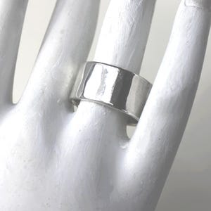 Op de afbeelding: Een brede, zilverkleurige ring met een plat, rechthoekig ontwerp. De ring wordt om een vinger gedragen en het oppervlak reflecteert licht, waardoor highlights en schaduwen ontstaan. De ring is ongeveer 1,3 cm breed.