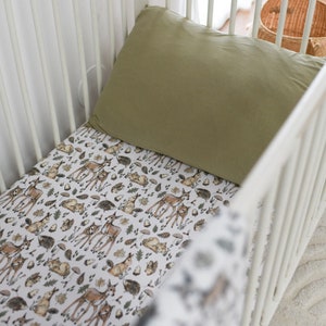 May include: A white cot with a green pillow and a fitted sheet featuring a woodland animal print. The sheet has a repeating pattern of deer, rabbits, hedgehogs, and leaves.
