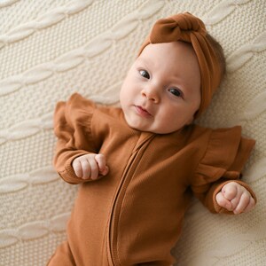 May include: A baby wearing a brown ribbed zippered onesie with ruffled sleeves and a brown knotted headband.