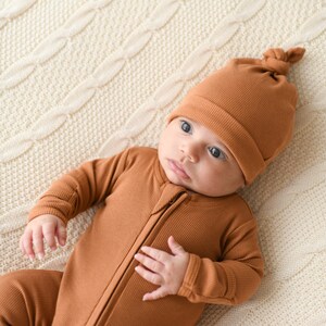 May include: A baby wearing a brown ribbed zippered onesie and a brown ribbed knotted hat.