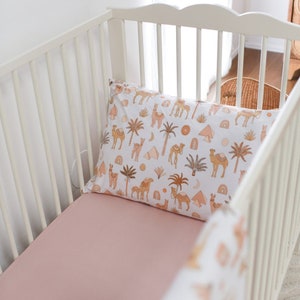 May include: A white crib with a pink fitted sheet and a pillowcase featuring a desert-themed pattern. The pillowcase has illustrations of camels, palm trees, pyramids, and rainbows in shades of brown and peach.