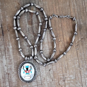 May include: Silver beaded necklace with a pendant featuring a Native American design. The pendant has a white background with a figure in red, blue, and black. The necklace is displayed on a wooden surface.